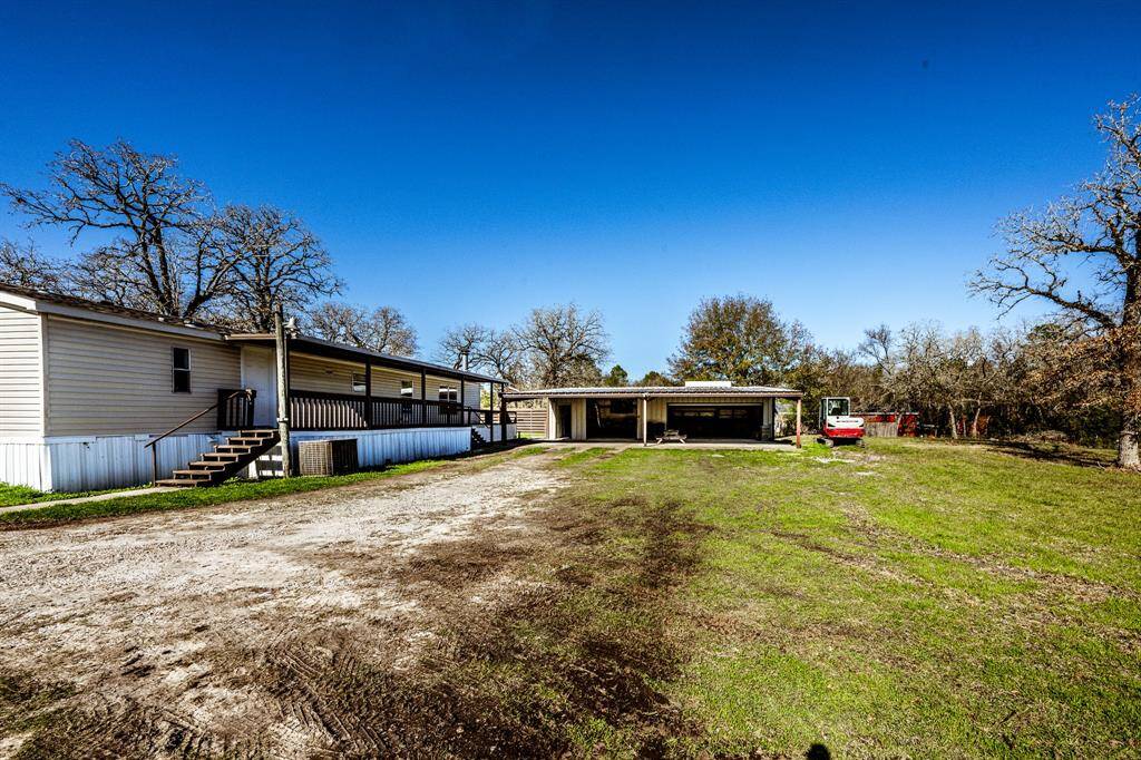 This photo showcases a spacious property with a mobile home featuring a large covered porch. The surrounding area includes open yard space, mature trees, and a detached carport, offering ample outdoor space and potential for customization.