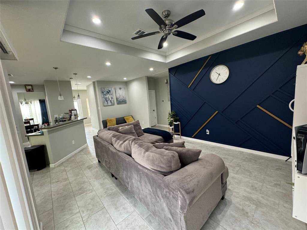 This modern living space features a spacious open layout with a stylish accent wall, recessed lighting, and a ceiling fan. The kitchen area includes a breakfast bar with pendant lights, and the room has a sleek, contemporary design with neutral tones.