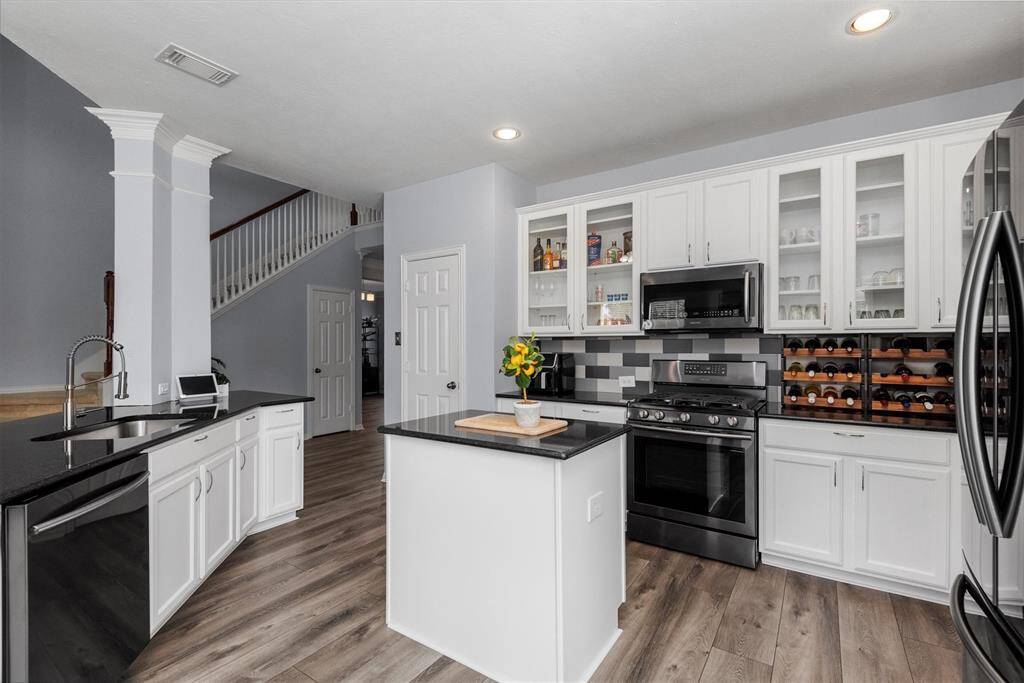 This is a spacious, modern kitchen featuring white cabinetry, stainless steel appliances, a center island with a sink, and an open layout leading to a staircase and additional rooms. The gray and white color scheme is complemented by wood-look flooring.