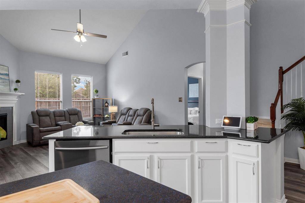 This is a spacious, open-plan living area featuring a modern kitchen with a central island and white cabinetry, looking out towards a comfortable living room with a cozy fireplace and ample natural light from windows. The space is finished with neutral colors and vinyl wood flooring, creating an inviting and contemporary atmosphere.