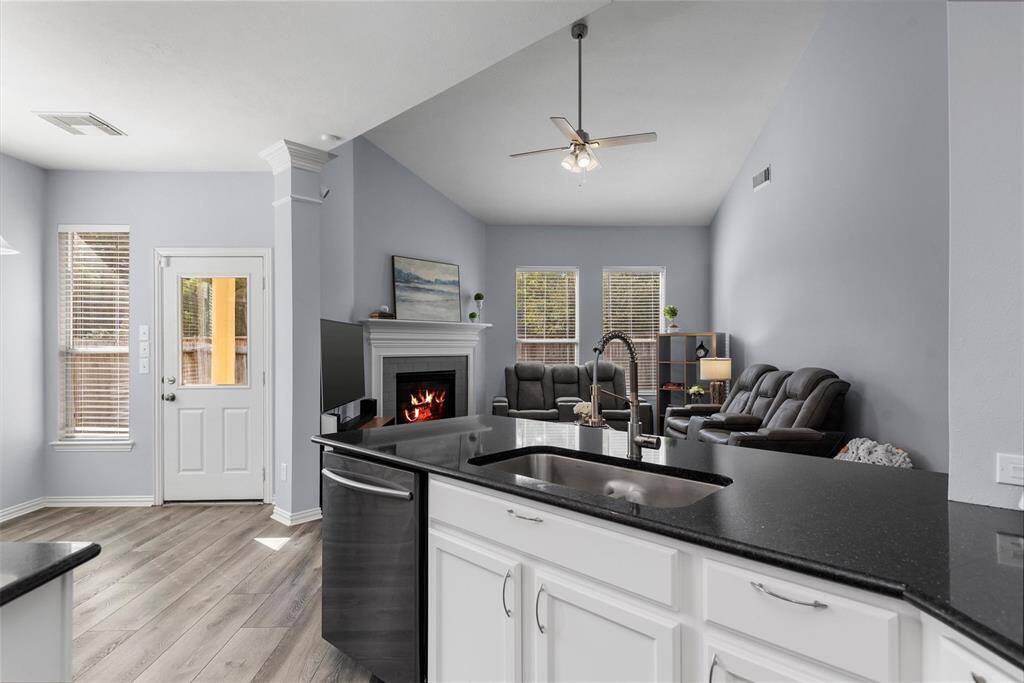 This is a modern, open-concept living space featuring a kitchen with a black countertop and white cabinets, stainless steel appliances, and a cozy living area with a fireplace. Natural light comes through the blinds of multiple windows, and there's a ceiling fan for airflow. The room has a neutral color palette with light gray walls and wood-look flooring.