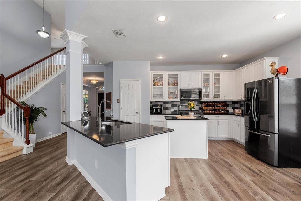 This is a spacious, modern kitchen with white cabinetry, stainless steel appliances, and a central island with a sink. The room features vinyl wood flooring and opens up to a staircase leading to the second level.
