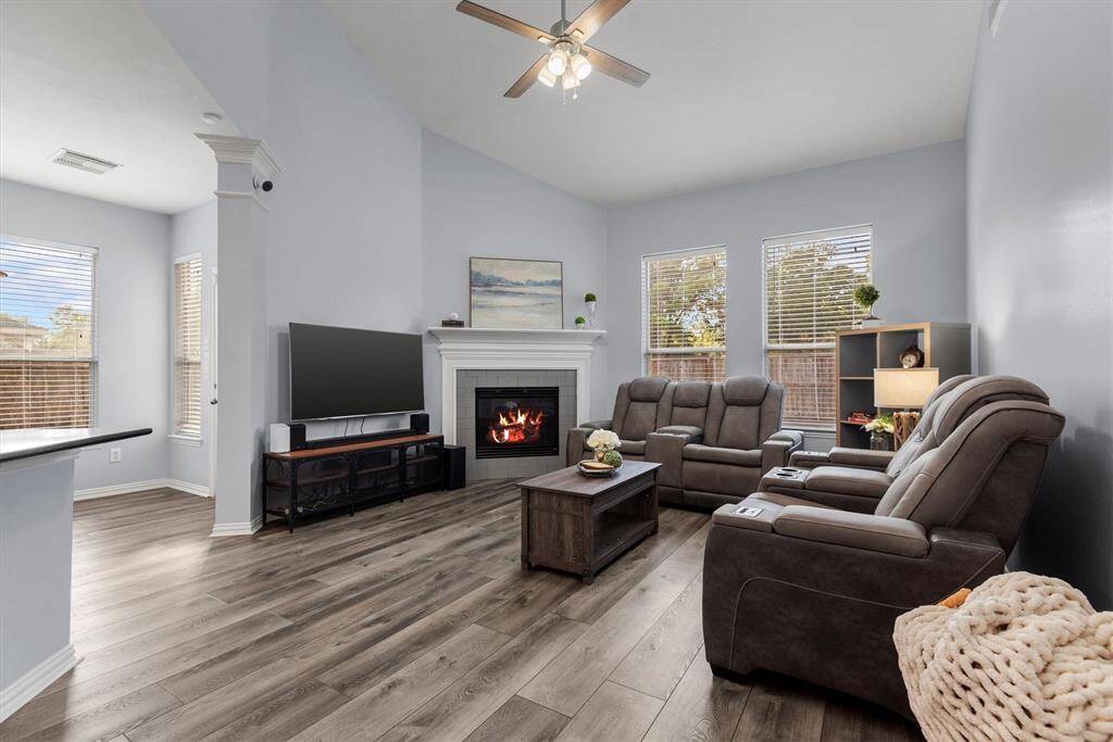 This is a cozy, well-lit living room featuring a vaulted ceiling with a ceiling fan, light gray walls, large windows with blinds, a classic white fireplace, and durable wood-look flooring.