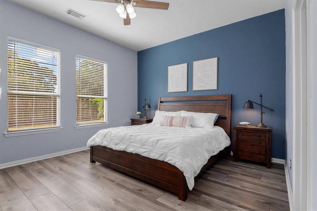This is a spacious bedroom featuring a calming blue accent wall, modern ceiling fan, two large windows for ample natural light, and stylish wood-look flooring.
