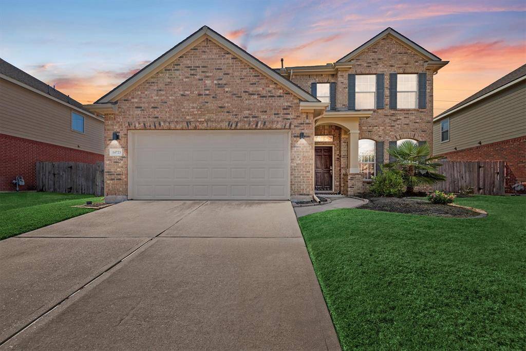 This is a two-story brick home featuring a two-car garage and a manicured lawn, set against a beautiful sunset sky. The house has a welcoming entrance with an arched doorway.