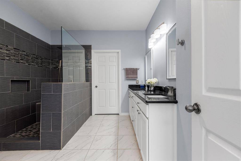 This is a modern bathroom featuring a walk-in shower with dark tile walls, a glass partition, a white vanity with a black countertop, and a large mirror with overhead lighting. The space has a clean, contemporary feel with light blue walls and white floor tiles.