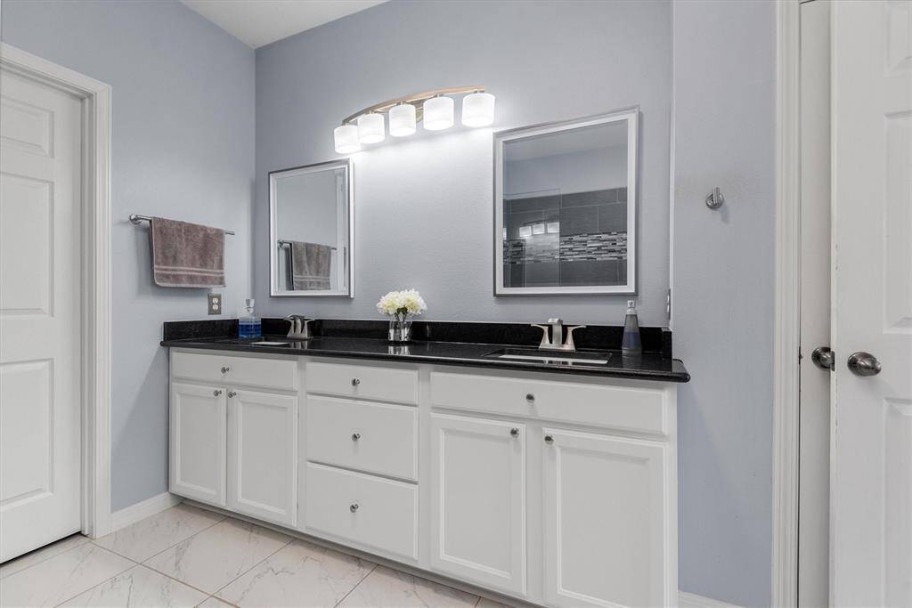 This is a clean, modern bathroom featuring a double vanity with white cabinetry and dark countertops, two mirrors, and a bright lighting fixture above. The walls are painted a soft blue, and the floor is tiled with a light marble design.