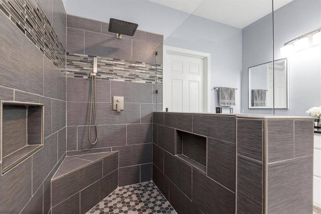 This is a modern bathroom featuring a walk-in shower with dark gray tiling, a rain shower head, and a detachable wand. There are built-in shelves within the shower area. The floor has hexagonal tiles, and the space is complemented by a light blue wall color, a white door, and a double vanity with mirrors.