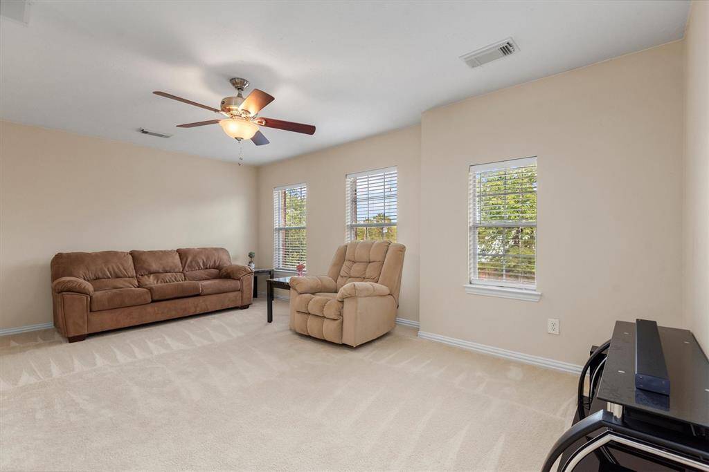 This is a well-lit, game room featuring neutral-painted walls, a plush carpet, a ceiling fan with a light fixture, and two windows that allow for natural light.