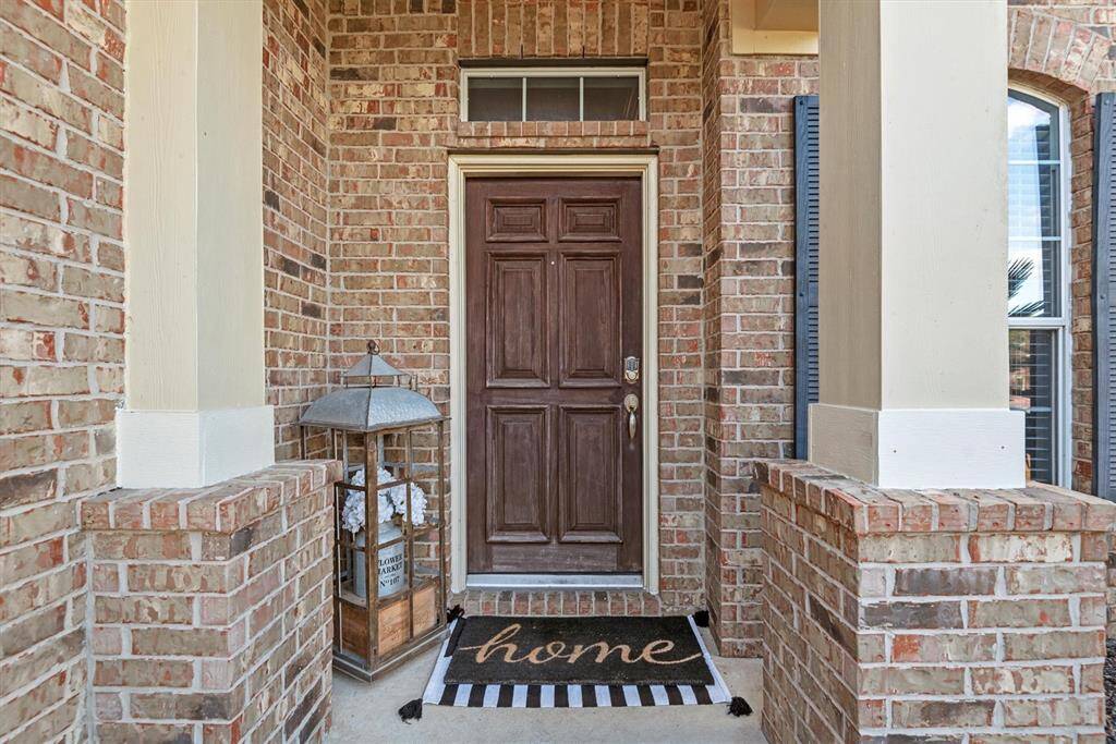 This photo shows a welcoming entryway with a traditional wood-stained front door framed by brick walls, accented with cream-colored trim and columns.