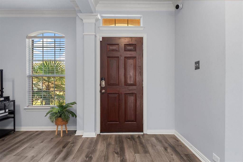 This is a bright entryway featuring a modern wood-look floor, a dark wood front door with glass transom above, crisp white trim, and a large window with plantation shutters that offer a view of greenery outside. The space is clean and neutral, ready for personalization.