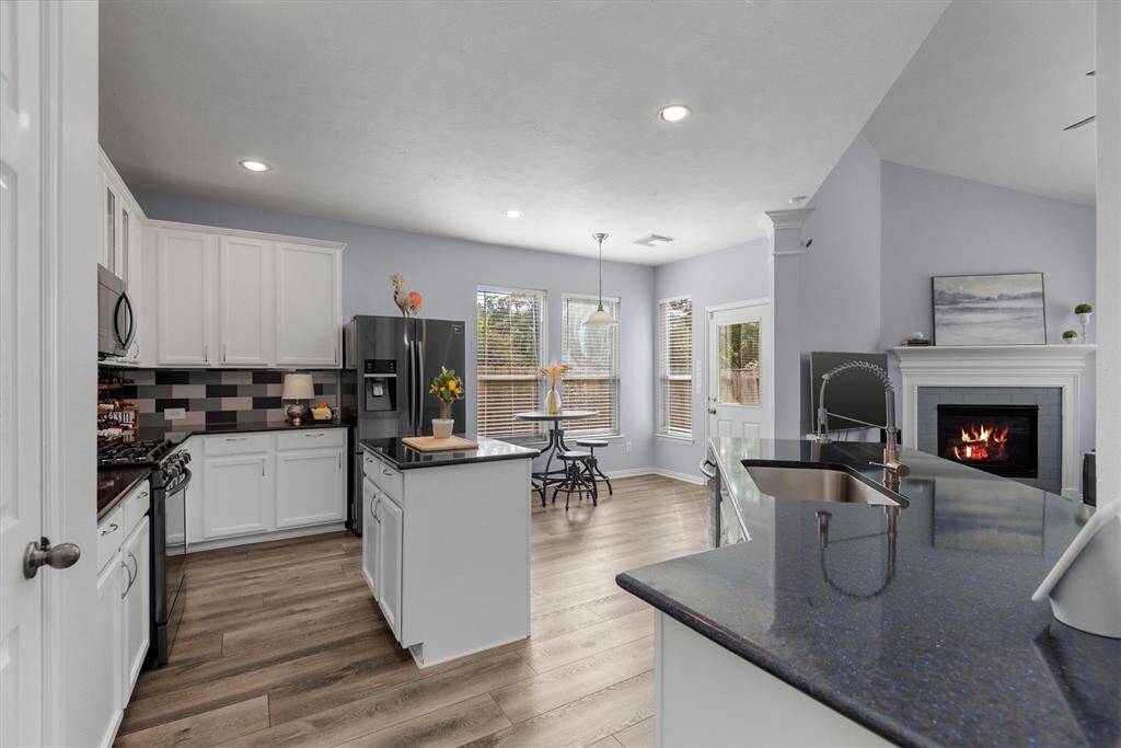 This is a modern, open-concept kitchen and living space featuring white cabinetry, stainless steel appliances, a stylish backsplash, and a central island with a sink. The area is complete with wood-like flooring, recessed lighting, and a cozy fireplace, all under a vaulted ceiling that enhances the room's spacious feel. Large windows allow for ample natural light.
