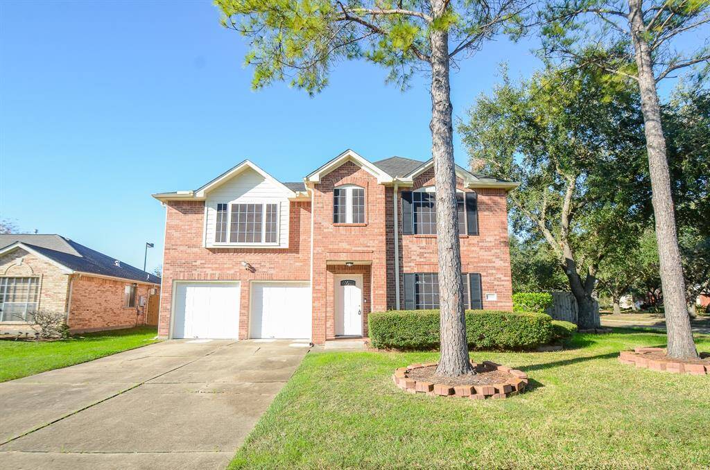 This photo features a two-story brick home with a double garage, large windows, and a well-maintained front yard with mature trees. The driveway is spacious, and the house is set in a suburban neighborhood.