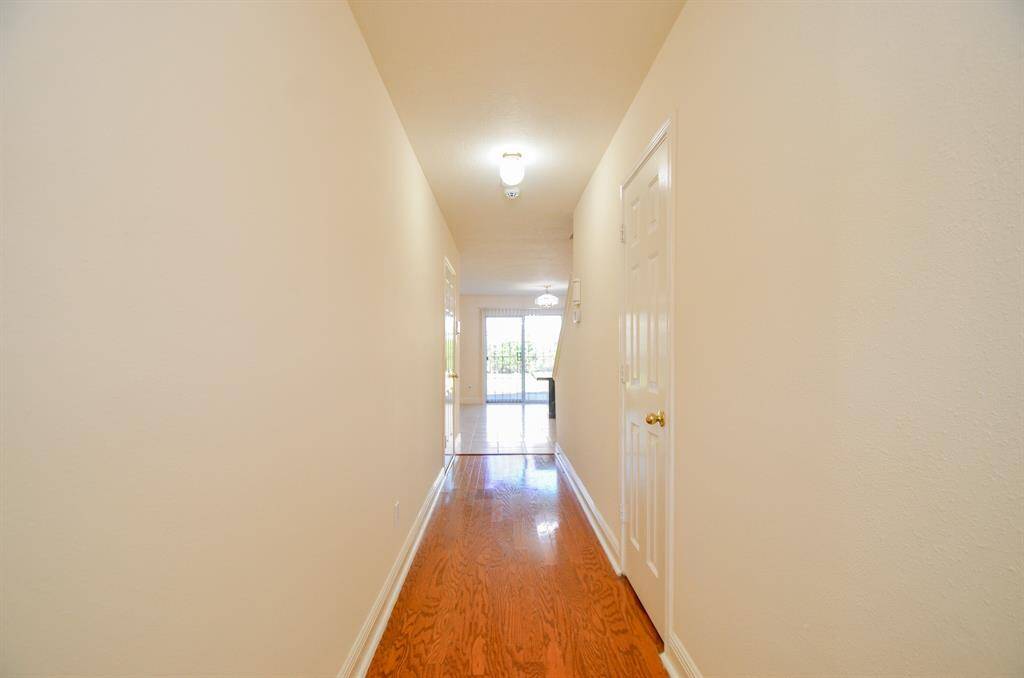 This photo shows a bright, long hallway with hardwood floors, neutral walls, and overhead lighting, leading to a room with large windows.