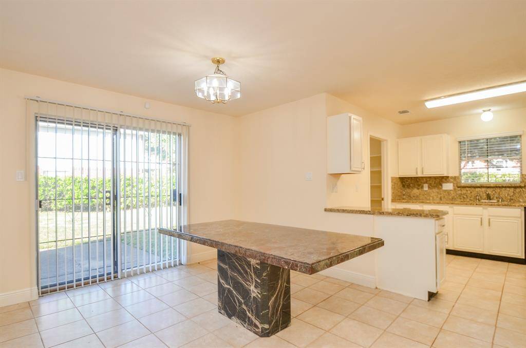 Bright dining area with a marble table, tiled floor, and sliding glass doors leading to a backyard. Adjacent kitchen features white cabinetry and granite countertops.