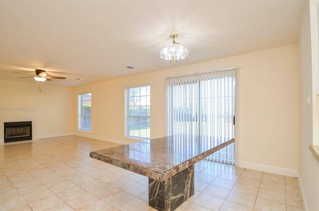 This spacious living area features a tiled floor, abundant natural light from large windows, a ceiling fan, and a cozy fireplace. It includes a marble dining table and sliding glass doors leading to the backyard, creating an inviting and functional space.
