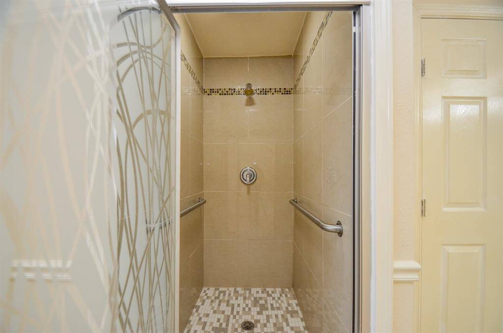 This photo showcases a modern shower with beige tile walls, decorative accents, and a sliding glass door with a stylish pattern. The shower features a built-in grab bar for safety and mosaic tile flooring.