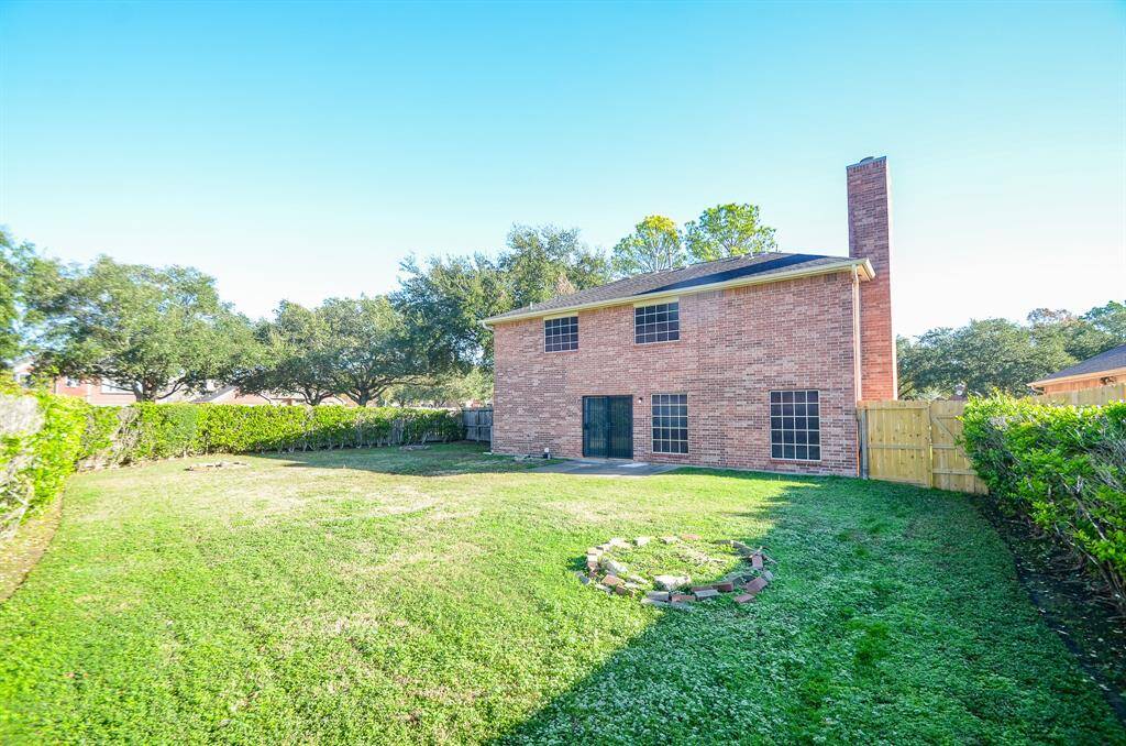 Spacious backyard with a well-maintained lawn, bordered by hedges for privacy. The two-story brick house features large windows and a patio area, perfect for outdoor activities.
