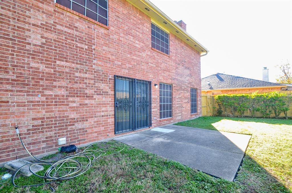 This photo shows the back of a two-story brick house with large windows and a patio area. The yard is grassy, with a hose reel on the ground and a wooden fence with shrubbery lining one side.
