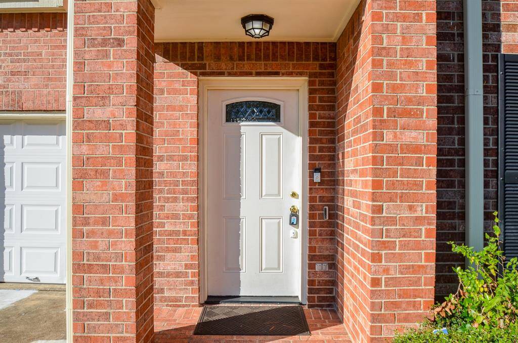 Charming front entry with a classic red brick exterior and a well-lit covered porch. Features a solid white door with decorative glass detail, adjacent to a garage. Perfect for welcoming guests.