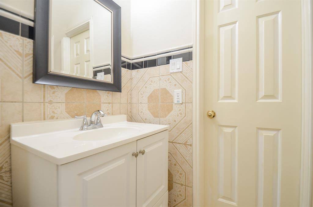 This photo showcases a compact bathroom with a modern vanity, featuring a sleek white sink and a stylish framed mirror. The walls are adorned with decorative tiles, and there's a light-colored door with a brass handle.