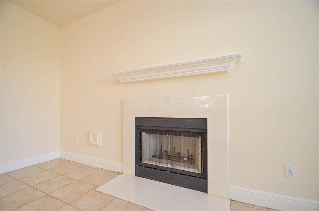 The photo features a cozy corner fireplace with a sleek black insert, complemented by a light marble surround and a simple white mantel. The room has neutral-toned tiled flooring and light-colored walls, creating a warm and inviting atmosphere.
