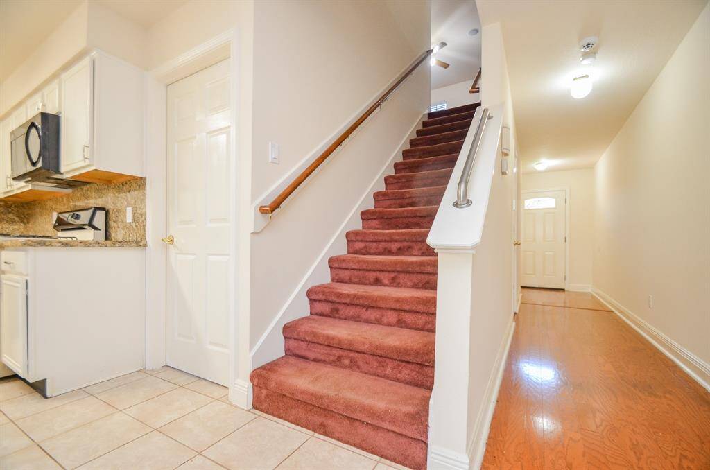 This photo shows an inviting entryway with red carpeted stairs, leading to the upper floor. To the left, there's a glimpse of the kitchen with light cabinetry and granite countertops. The hallway features polished wood flooring and a bright front door.