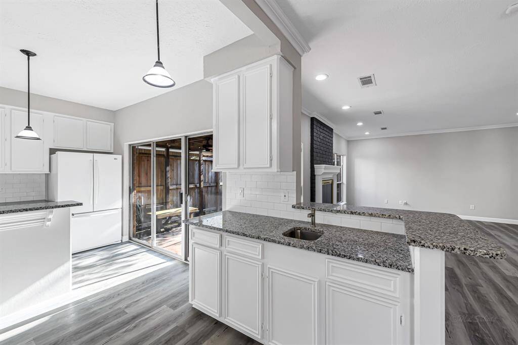 This photo showcases a modern kitchen with white cabinetry, granite countertops, and a spacious open layout. The kitchen leads to a living area with a fireplace, and there's a large sliding glass door that opens to an outdoor space. The flooring is a sleek, contemporary wood finish, enhancing the airy and bright atmosphere.