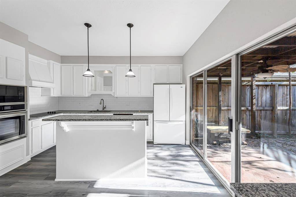 This modern kitchen features sleek white cabinetry, granite countertops, and stainless steel appliances. It is well-lit with pendant lights and natural light from large sliding glass doors that open to a private patio area.