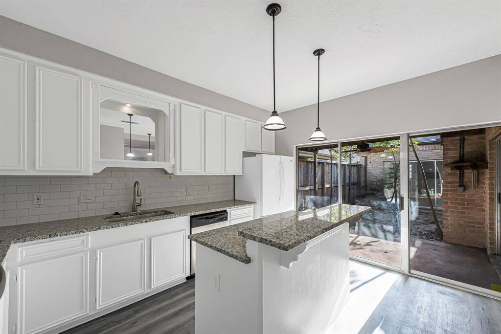 This kitchen features modern white cabinetry, granite countertops, and a large island. It’s well-lit with pendant lights and has sliding glass doors leading to a patio, offering plenty of natural light.