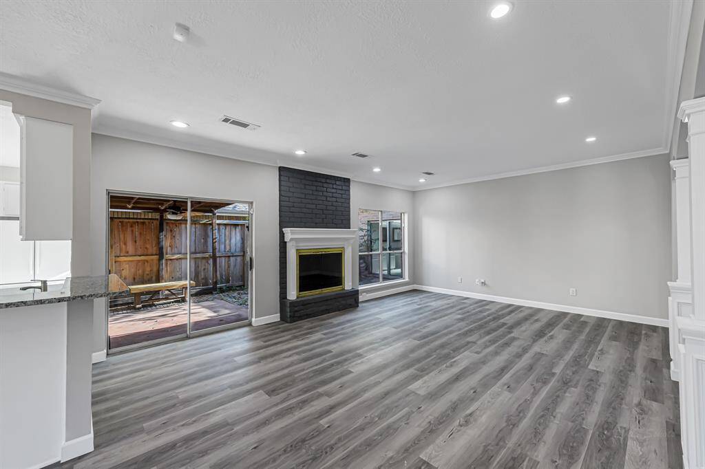 This spacious living room features modern gray flooring, a cozy fireplace with a sleek dark accent, and ample natural light from large sliding glass doors leading to an outdoor patio area. The open layout connects seamlessly to the kitchen, enhancing the home's inviting atmosphere.
