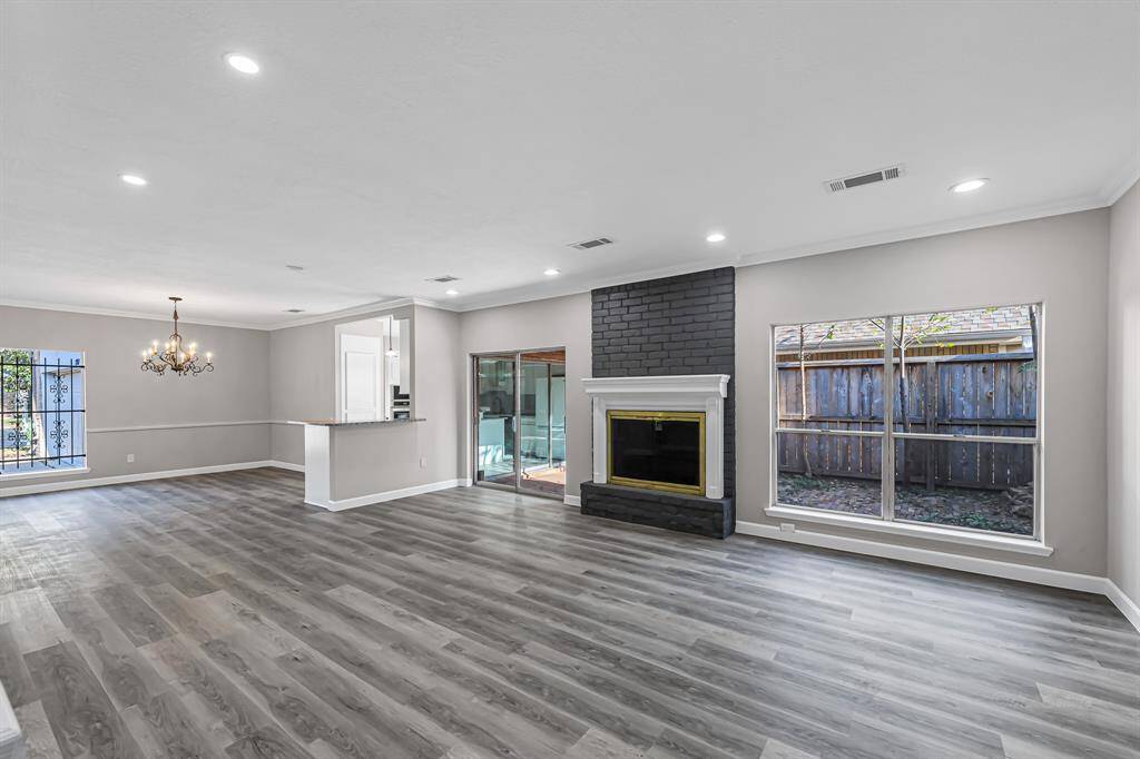 This spacious living area features modern gray flooring, a sleek dark painted fireplace, large windows for natural light, and an open layout with a dining area and kitchen pass-through. Perfect for entertaining and cozy nights in.