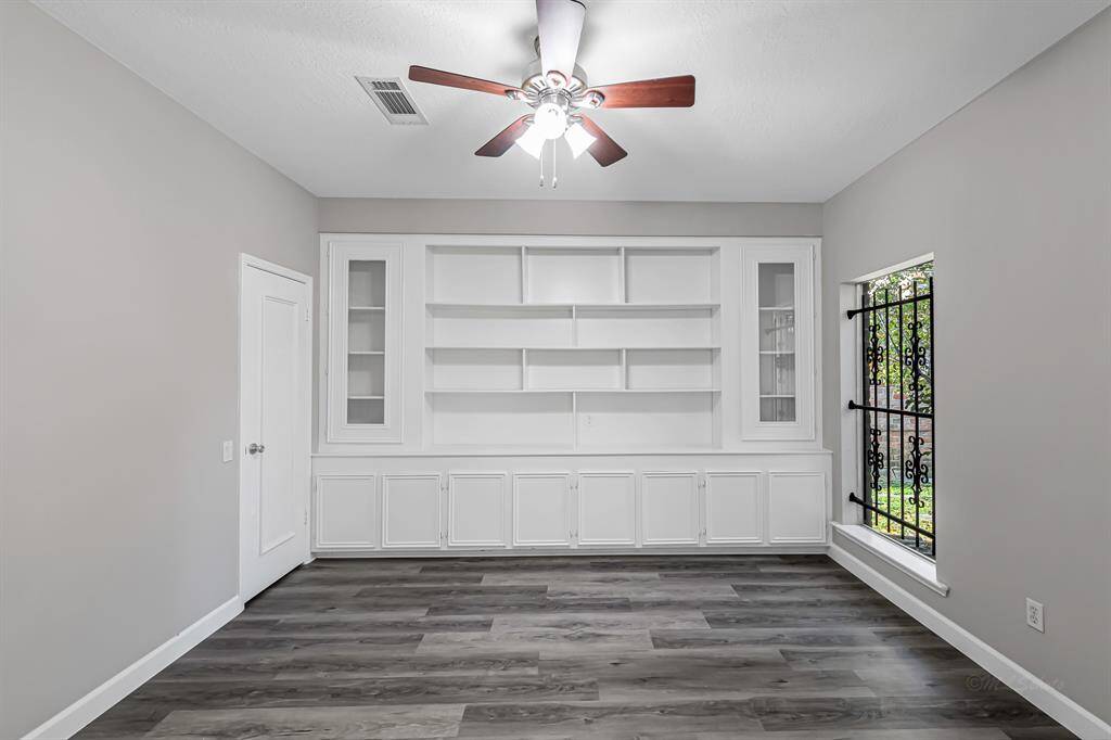 This room features sleek wood-look flooring, a ceiling fan, and a large built-in shelving unit. A window with decorative ironwork provides natural light, complementing the neutral walls. Ideal for a study or library space.