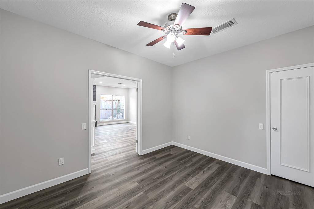 This photo shows a bright, open room with light gray walls and wood-look flooring. It features a ceiling fan and a large doorway leading to another room with a big window, providing plenty of natural light.