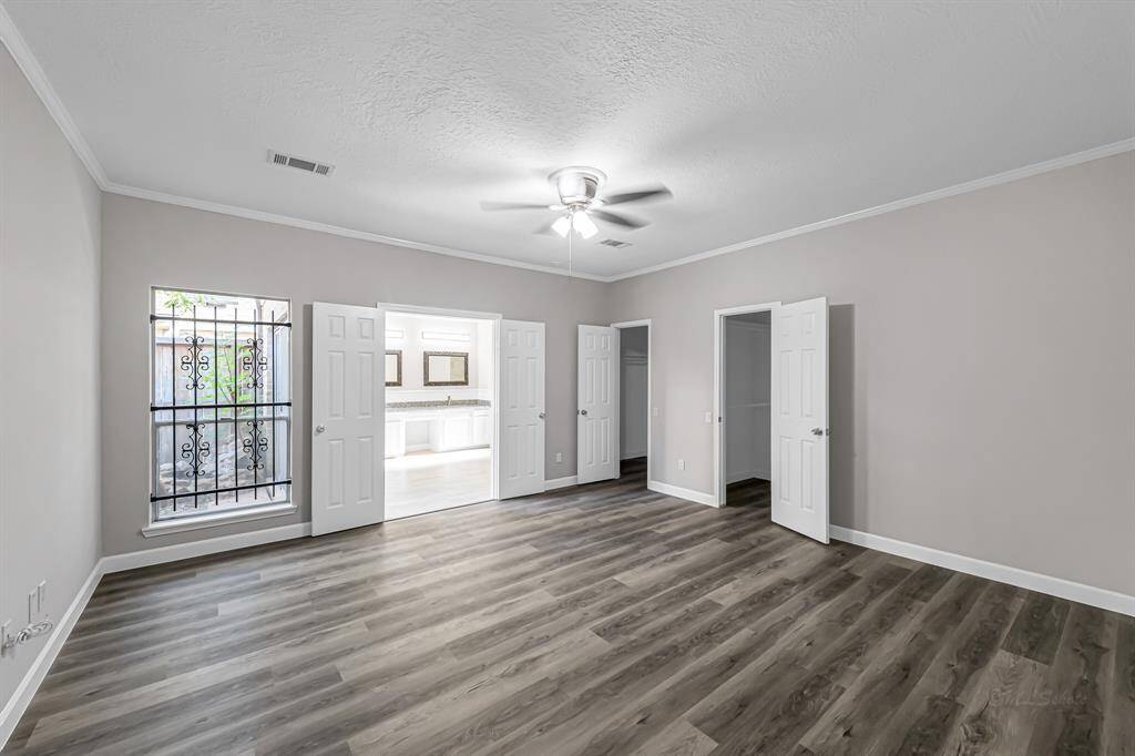 This room features sleek wood-style flooring, neutral walls, and a ceiling fan. It has ample natural light from a large window with decorative bars and offers access to a spacious bathroom and closet through double doors.