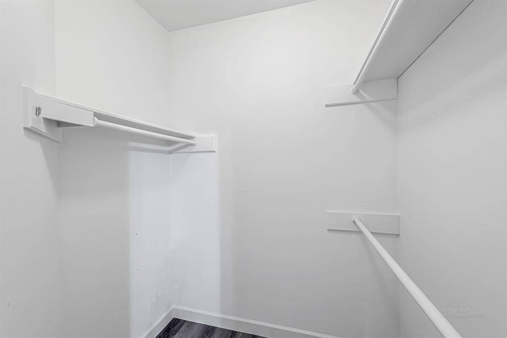 This photo shows a spacious walk-in closet with white walls and multiple hanging rods, offering ample storage space. The floor is finished with dark wood.
