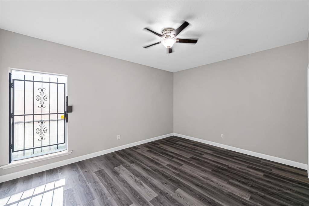 This room features light gray walls, dark wood-style flooring, and a ceiling fan. A window with decorative wrought iron bars allows natural light to fill the space.