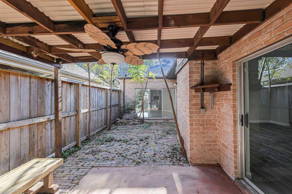 This photo shows a cozy outdoor covered patio with a wooden trim and ceiling fan, surrounded by brick walls and a wooden fence. It features sliding glass doors and a rustic bench, perfect for relaxing or entertaining.