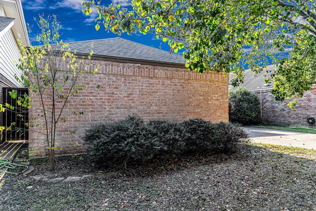 This is a view of the front brick exterior with a small garden area featuring shrubs and a small tree, adjacent to a paved driveway. The area is partially shaded by a larger tree, offering a cozy outdoor space.