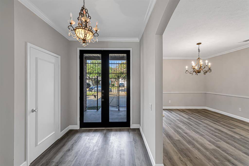This photo showcases a bright entryway with elegant chandeliers, French doors, and wood-style flooring, leading into a spacious living area with crown molding.