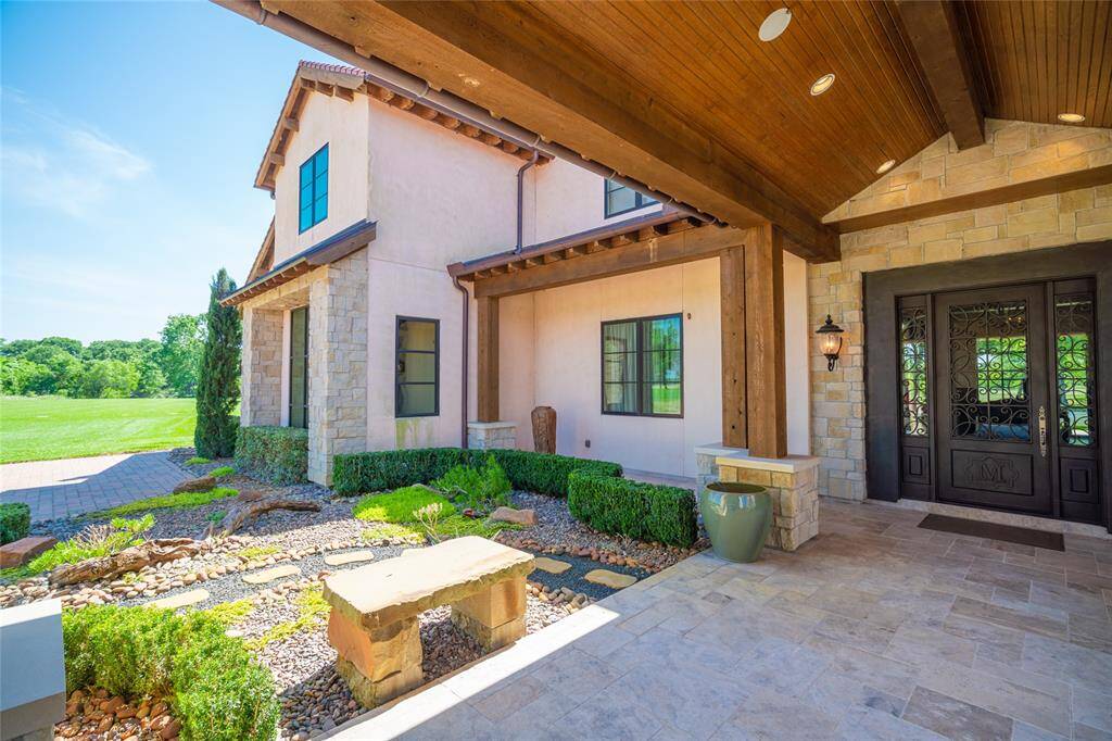 A grand motor court ENTRY to the Main House with its covered stained wood planked pitched portico ceiling with recessed lighting, hanging chandelier, stone pillars, stone tile flooring, handsome glass/wood front door/flanking windows with decorative iron design, noteworthy light sconces, outdoor speakers - all surrounded by well maintained rock gardens and stone benches.