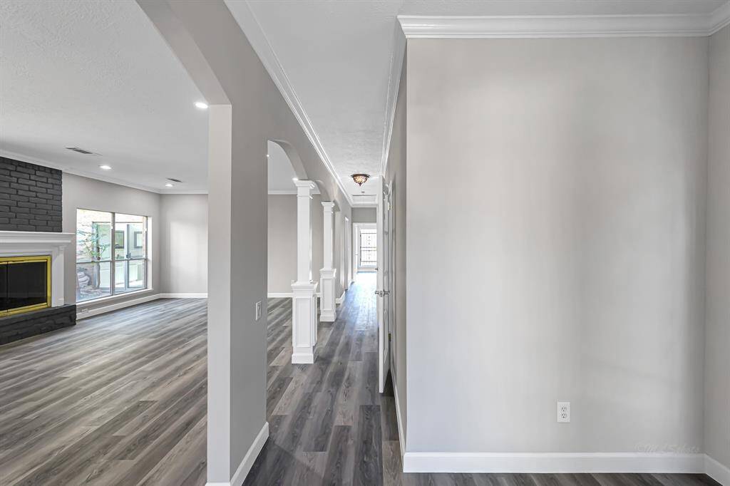 This photo showcases a modern interior with an open floor plan, featuring sleek wood flooring and neutral walls. The space is well-lit with natural light from large windows, and includes a stylish black brick fireplace. Decorative columns add elegance to the hallway.
