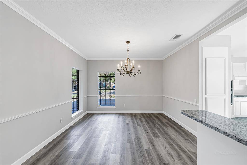 This photo showcases a bright dining area with stylish wood-look flooring, a classic chandelier, and ample natural light from two windows. The space features neutral walls and elegant crown molding, adjacent to an open kitchen with granite countertops.