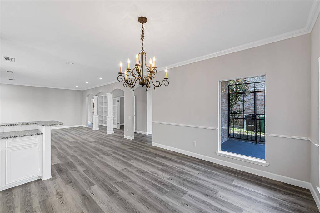 This photo showcases a spacious, open-concept living area with elegant wood flooring, a large window for natural light, and a classic chandelier. The room includes a view of wet bar counter with granite countertops, creating a perfect blend of style and functionality.
