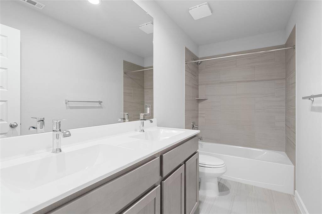 Secondary bath features tile flooring, bath/shower combo with tile surround, stained wood cabinets, beautiful light countertops with dual vanities, mirror, dark, sleek fixtures and modern finishes.