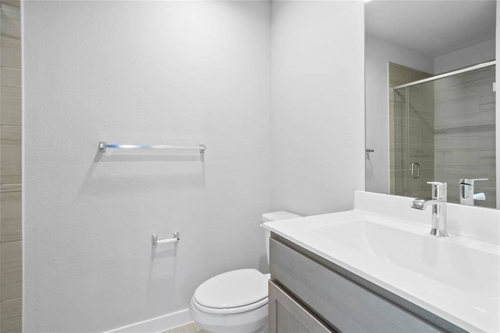 Secondary bath features tile flooring, walk-in shower with tile surround, stained wood cabinets, beautiful light countertops, mirror, dark, sleek fixtures and modern finishes.