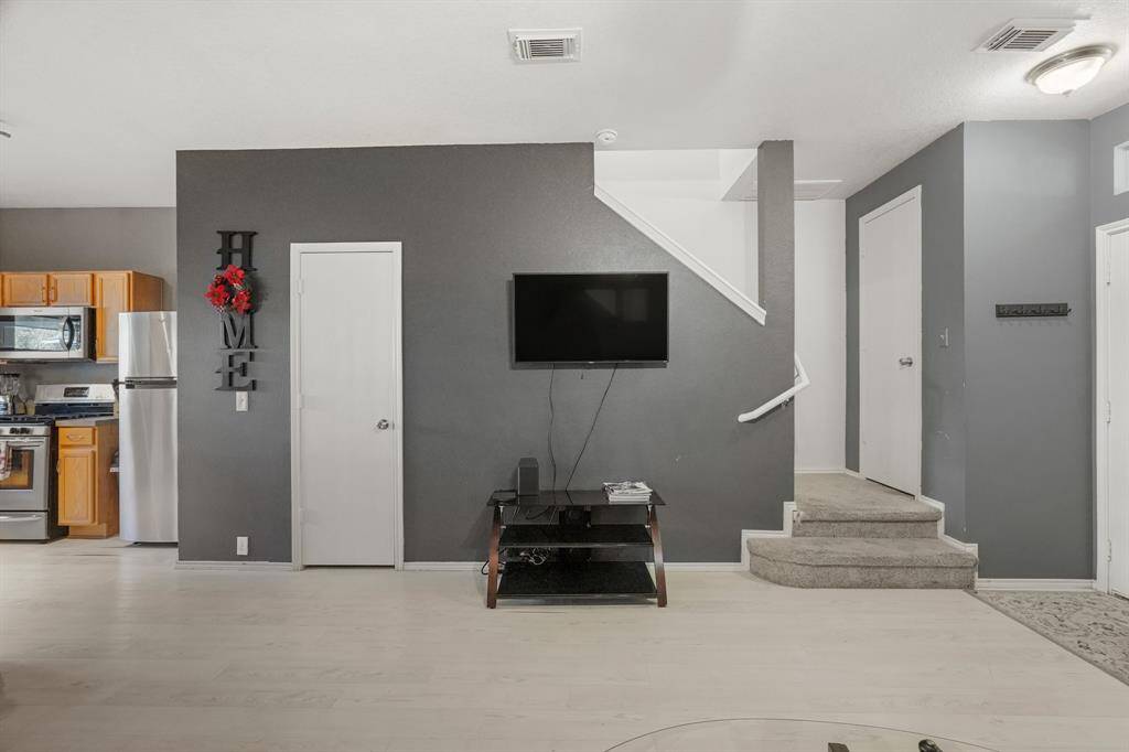 This photo shows a modern living area with light flooring, a wall-mounted TV, and a staircase leading upstairs. The kitchen area features stainless steel appliances and wooden cabinetry. The space is well-lit, with a neutral color palette and decorative accents.