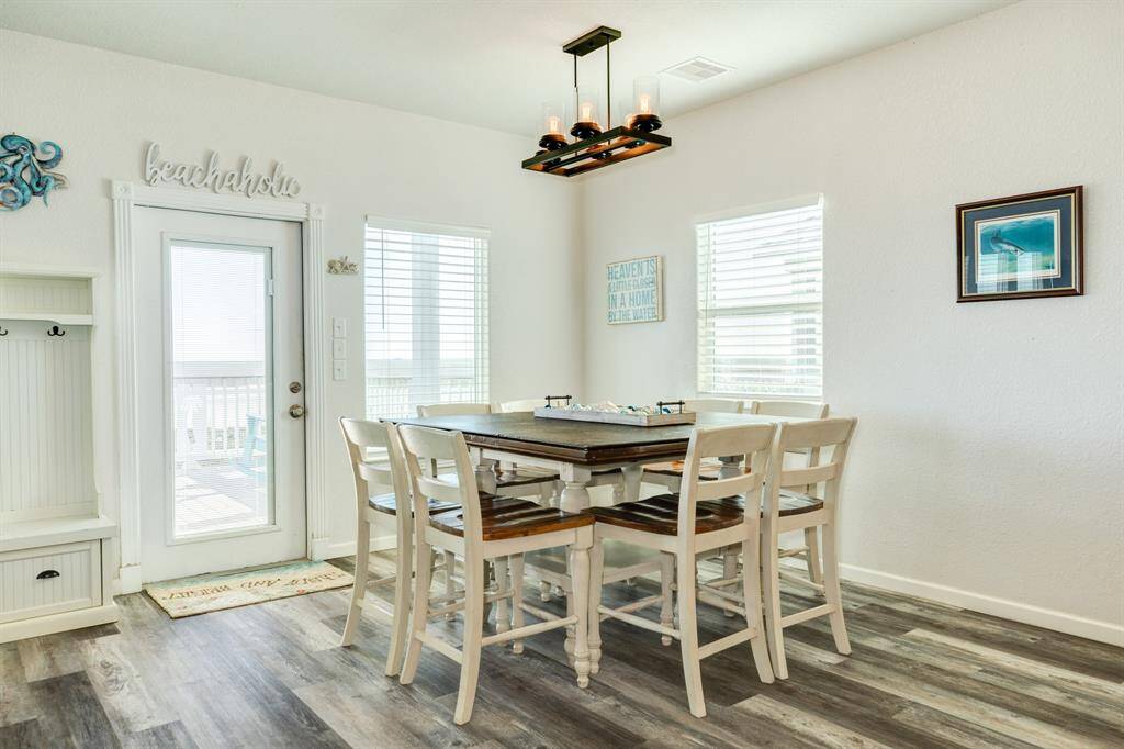 Bright dining area with a coastal theme, featuring a wooden table and chairs, modern light fixture, and access to a deck through a glass door. Light-colored walls and flooring enhance the airy feel.