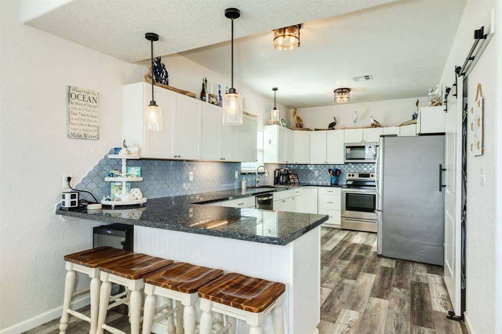 This modern kitchen features sleek white cabinetry, stainless steel finish appliances, and a spacious granite countertop with bar seating. The stylish backsplash and wood-look flooring add warmth and character to the space, enhanced by pendant and ceiling lighting.