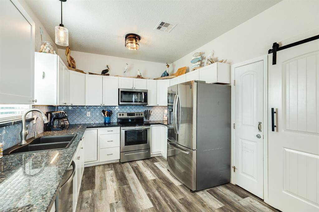 Modern kitchen with stainless steel finish appliances, white cabinetry, and a stylish blue backsplash. Features granite countertops, wood-look flooring, and a sliding barn door for a cozy, contemporary feel.